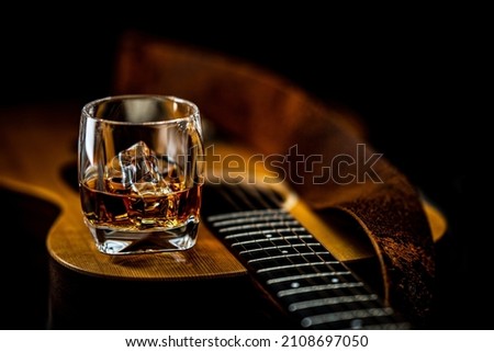 An acoustic guitar lays on it's back while a glass of whiskey with ice rests on it's top Royalty-Free Stock Photo #2108697050