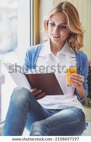 Beautiful young woman holding a glass of juice, reading a book and smiling while sitting on the window sill