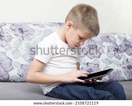 The boy sits on the couch and looks at the tablet