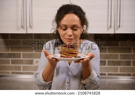 Charming woman holding a plate of delicious homemade freshly baked pancakes garnished with wild berries. Beautiful young dark-haired curly housewife with cooked pancakes for Shrovetide. Shrove Tuesday Royalty-Free Stock Photo #2108741828