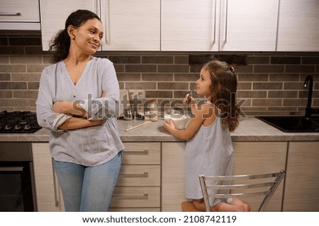 Beautiful smiling woman, happy loving mother leaning at kitchen countertop and admiring her adorable daughter cute baby girl mixing ingredients in a glass bowl while learning kneading pancakes dough