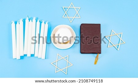 One large donuts with powder and cream in a saucer, nine white candles, a bible and three wooden stars of David lie on a blue background, flat lay close-up. Hanukkah Celebration Concept.