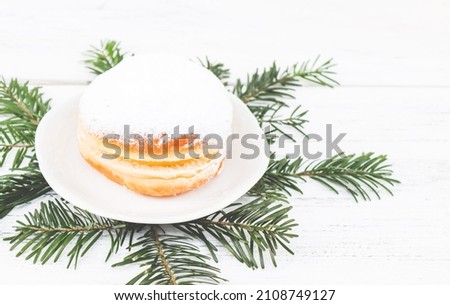 A donut with powdered sugar and cream in a saucer with fir branches lie on a white wooden table, close-up side view. Hanukkah celebration concept.