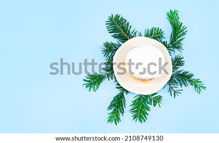 One large donuts with powder and cream in a saucer on fir branches lie on the right against a blue background with copy space on the left, flat lay close-up. Hanukkah Celebration Concept.