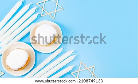 Two large donuts with powder and cream in saucers, nine white candles and two wooden david stars lie on the left against a blue background with copy space, flat lay . Hanukkah Celebration Concept.
