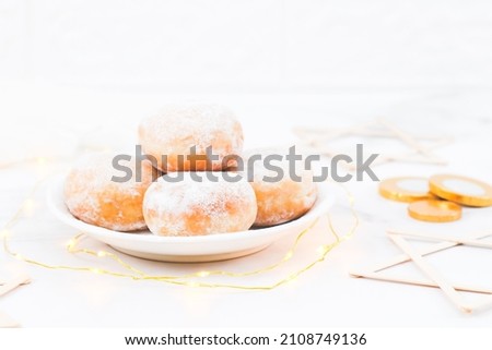 Donut with powdered sugar and cream in a saucer with a garland, coins and wooden Star of David lie on a white wooden table, close-up side view. Hanukkah celebration concept.