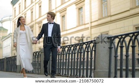 Beautiful newlyweds walking around city. Action. Happy and elegant newlyweds are walking along pavement of city canal. Walking newlyweds in city Royalty-Free Stock Photo #2108750072