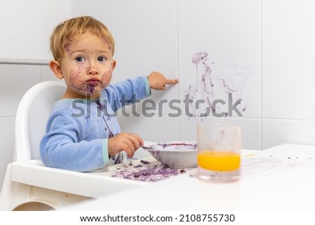 blond boy in his high chair shows the wall that he has stained with his breakfast Royalty-Free Stock Photo #2108755730