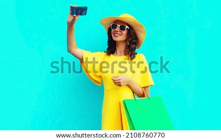 Beautiful happy smiling woman taking selfie by smartphone with shopping bags wearing yellow dress, summer straw hat on colorful blue background