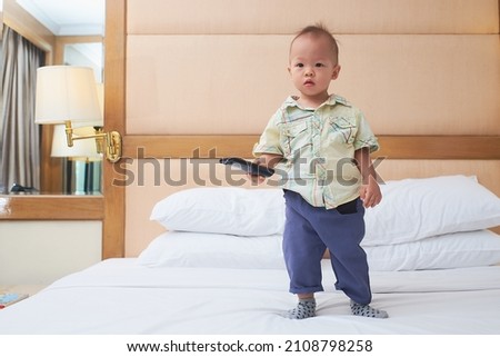 Cute little Asian 1 year old toddler baby boy child standing in bed holding the tv remote control and watching television in bedroom at home, Toddlers Screen Time, Early Brain Development concept