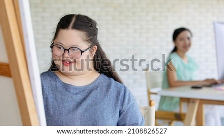 Portrait shot of Asian happy lovely mother standing smiling with young chubby down syndrome autistic autism little daughter wearing eyeglasses holding paintbrush drawing painting on canvas easel.