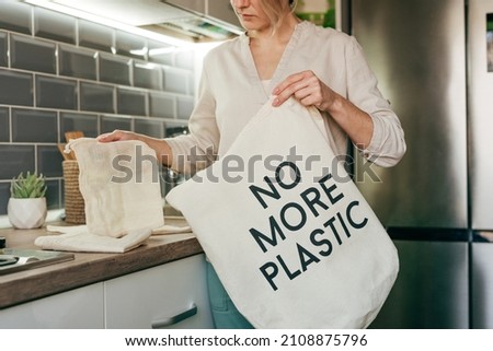 Young woman standing in the kitchen putting reusable cotton produce bags into the shopper bag with inscription NO MORE PLASTIC. Zero waste concept.