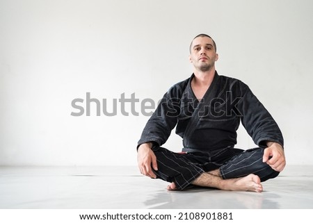 Front view of adult male athlete bjj brazilian jiu jistu black belt sitting in front of white wall in kimono gi with copy space looking to the camera Royalty-Free Stock Photo #2108901881