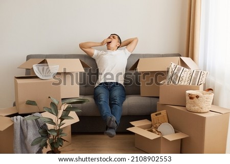 Indoor shot of exhausted sleepless man wearing white T-shirt and jeans sitting on cough surrounded with pile of packages and yawning, being tired and sleepy with relocating.