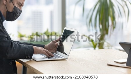 A businessman in black dress wearing face mask is using laptop to communicate with friends and colleagues. Attractive male manger is sitting in green office space with cityscape background 