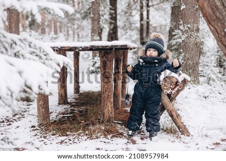 Child walking in snowy spruce forest. Little kid boy having fun outdoors in winter nature. Christmas holiday. Cute toddler boy in blue overalls and knitted scarf and cap playing in park.