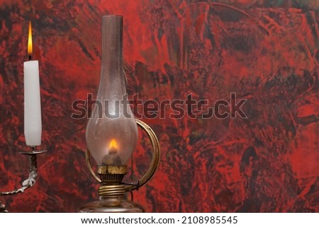 A fragment of an old, glowing oil lamp and a lit white candle against a red and black wall.
