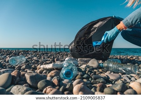 Close-up of female hands collecting plastic, rubber garbage in a black garbage bag on a pebble beach against the background of the sea with copy space. Environmental pollution on the Black Sea coast.