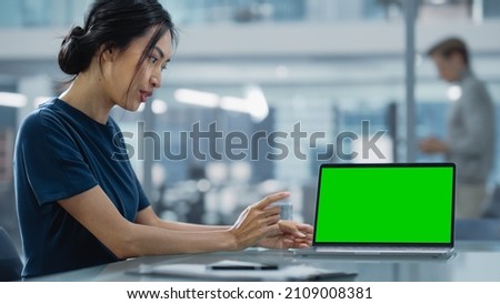 Multiethnic Office Meeting Room: Creative Asian Entrepreneur Female Use Laptop and Pointing on Green Screen Mock Up Display.