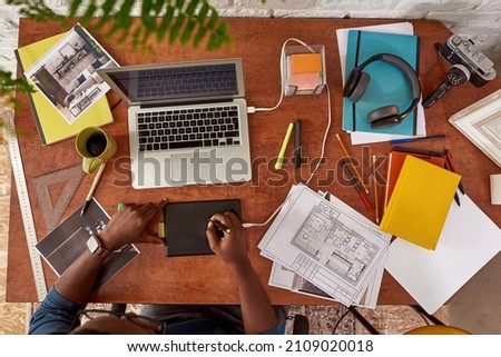 Top view of male architect drawing building plan on graphics tablet and watching on laptop computer. Partial image of young black millennial guy working at home. Concept of freelance and remote work
