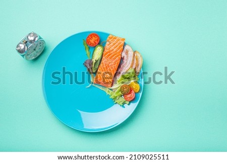 Intermittent fasting low carb hight fats diet concept flat lay, healthy food salmon fish, bacon meat, vegetables and salad on blue plate and clock alarm on blue background top view, space for text Royalty-Free Stock Photo #2109025511