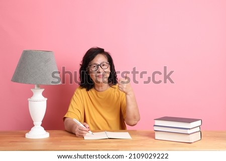 The senior Asian woman reading book sitting at the desk with the pink background.
