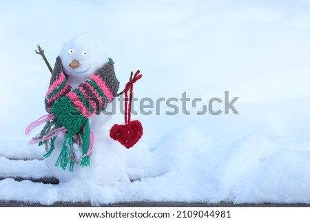 happy snowman in a scarf holds a symbol of a heart knitted from threads on the background of winter snowdrifts. warming valentine