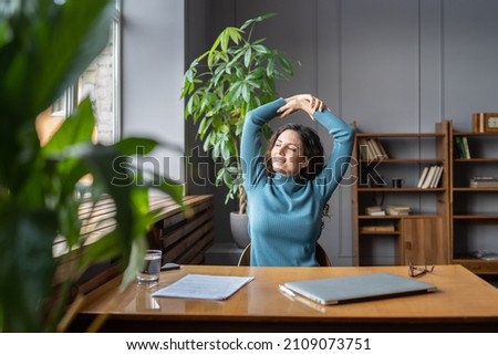 Smiling young female employee relaxing at workplace, sitting in chair stretching arms, doing gymnastic in office, looking in window with pleased face expression, feeling satisfied with work results Royalty-Free Stock Photo #2109073751