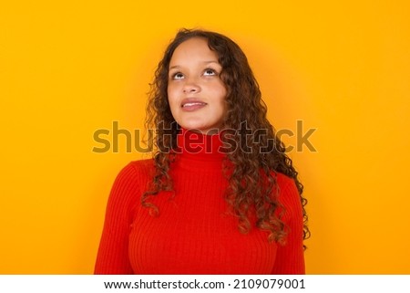 Portrait of mysterious Young beautiful girl wearing red sweater standing against yellow background looking up with enigmatic smile. Advertisement concept.