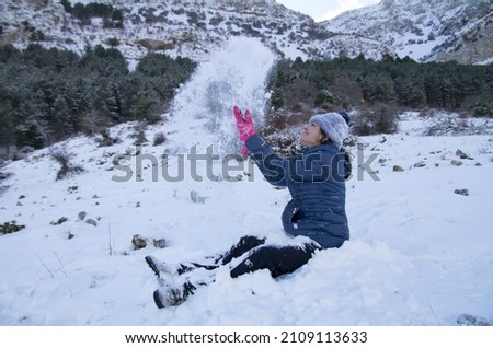 a happy brunette latin girl, sitting in the snow, throwing it up and laughing, wearing black pants, a padded coat, a blue wool hat and pink gloves