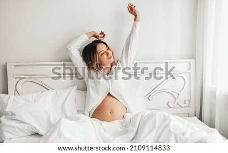 Tender pregnant female in nightwear sitting on bed and stretching with raised arms after awakening in morning Royalty-Free Stock Photo #2109114833