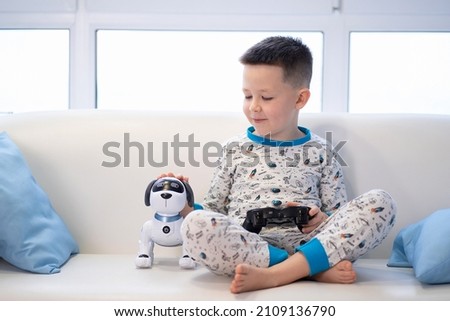 The boy sits on the couch in a hug, strokes the robot dog and plays video games Royalty-Free Stock Photo #2109136790