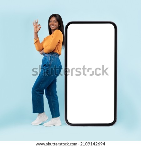 Mobile Offer. Happy black woman leaning on huge cellphone with blank white screen, showing ok gesture, recommending great new app or website offering free space for ad, mock up, full body length
