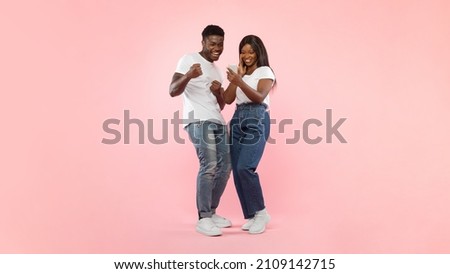 Portrait Of Joyful African American Couple Holding Sharing Smart Phone Shaking Fists In Joy And Excitement, Celebrating Online Win Standing Over Pink Studio Background. Yes, Great News Message Concept Royalty-Free Stock Photo #2109142715
