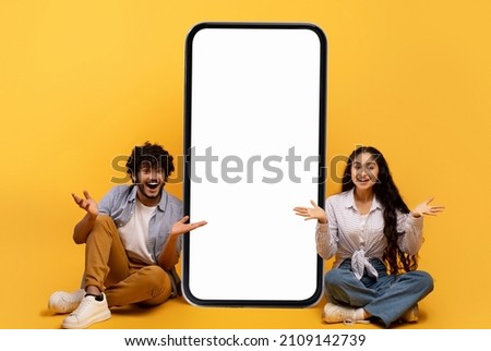 Surprised indian couple sitting near huge smartphone with blank screen over yellow studio background, demonstrating mockup for mobile app or website. Cellphone template