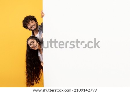 Excited indian couple standing behind white advertisement board and peeking out, showing empty space for your design or offer, posing over yellow studio background, free space Royalty-Free Stock Photo #2109142799