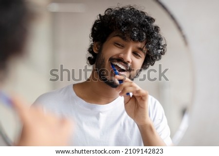 Oral care concept. Young indian man cleaning teeth with toothbrush, smiling to his reflection in mirror, doing toothcare hygiene routine in the morning in bathroom Royalty-Free Stock Photo #2109142823