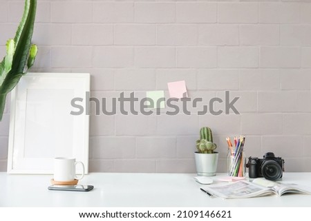 Stylish workplace with empty picture frame, cactus, camera and coffee cup on white table.