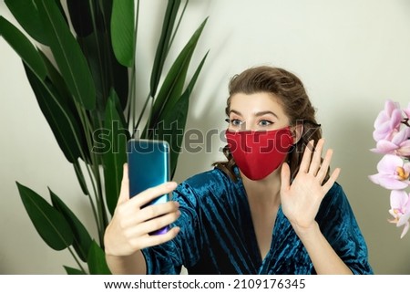 Young woman with medical protective mask on face dressed in pin-up style uses mobile phone. Woman talks with friend or family during lockdown at home.  Selective focus