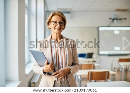 Smiling mature teacher holding laptop while standing in the classroom and looking at camera. Royalty-Free Stock Photo #2109199061