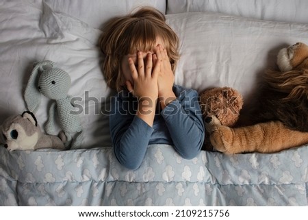 The child was scared before going to bed. Night terrors in a child. The kid covers his face with his hands in a fear. Children's experiences. Boy in the bed. View from above. Sad psychological state. Royalty-Free Stock Photo #2109215756