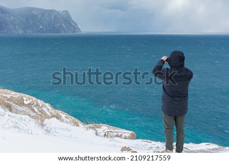 Man phone sea snow. A male tourist photographs a winter landscape. Top view of the blue sea and heavy snowfall. A fabulous winter landscape with mountains and turquoise water. Enjoy the beautiful view