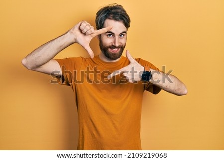 Caucasian man with beard wearing casual yellow t shirt smiling making frame with hands and fingers with happy face. creativity and photography concept. 