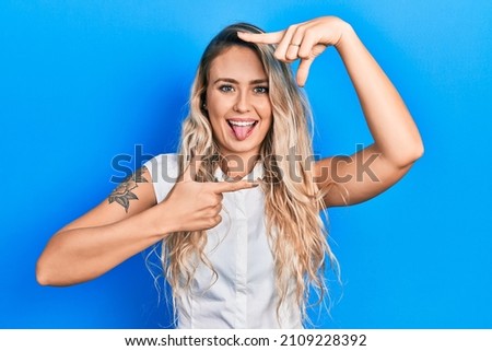 Beautiful young blonde woman doing picture frame gesture with hands sticking tongue out happy with funny expression. 