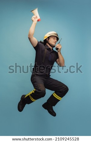 Giving directions. Portrait of young firefighter in uniform with walkie-talkie and megaphone jumping over blue studio background. Concept of different professions, unusual people and their emotions. Royalty-Free Stock Photo #2109245528