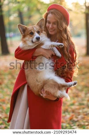 A girl with long red hair, in a red coat and beret gently hugs her corgi dog in the autumn park. Golden autumn, walking with a dog, friendship, dancing with a dog. beautiful autumn picture