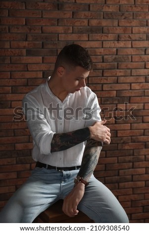 Young man with tattoos sitting on stool near brick wall