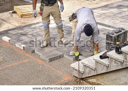 Construction workers wearing safety protective gear and working on high quality landscaping driveway pavement site. Contractors laying interlock patio with stone brick tile. Royalty-Free Stock Photo #2109320351