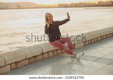 Beautiful Asian lady holding smartphone and taking picture of herself while sitting on waterfront on the backgroung of calm sea
