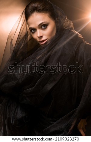 Attractive stylish female with beautiful makeup posing for the photo, looking at the camera. Gorgeous young adult woman wearing fashion black dress and veil, modern bride concept